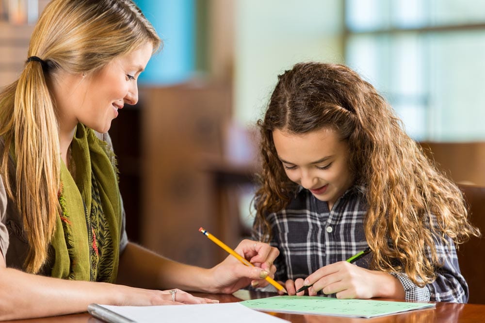 Elementary age Caucasian little girl with brown curly hair is working on math worksheet while doing homeschool assignment or homework with her mother. Mid adult Caucasian woman is teaching her young daughter in public library.