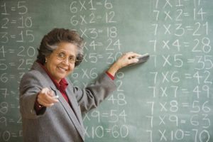 older teacher in front of chalkbaord with math problems smiling and pointing at camera