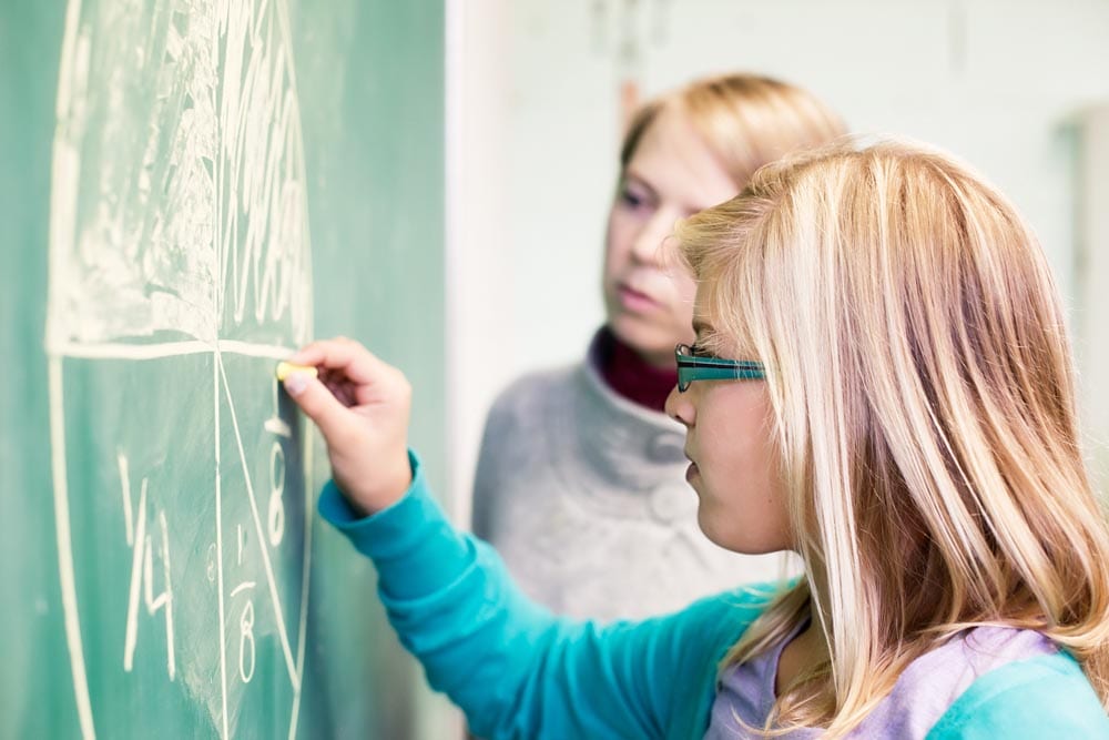 Preteen girl learning fraction by drawing a pie on the chalk board under the supervision of her teacher.