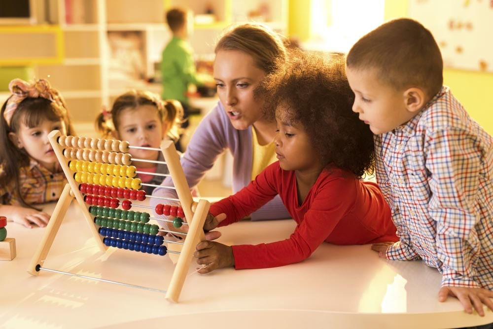 Group of kids and teacher using abacus at kindergarten. Focus is on African American little girl. There are children in the background.