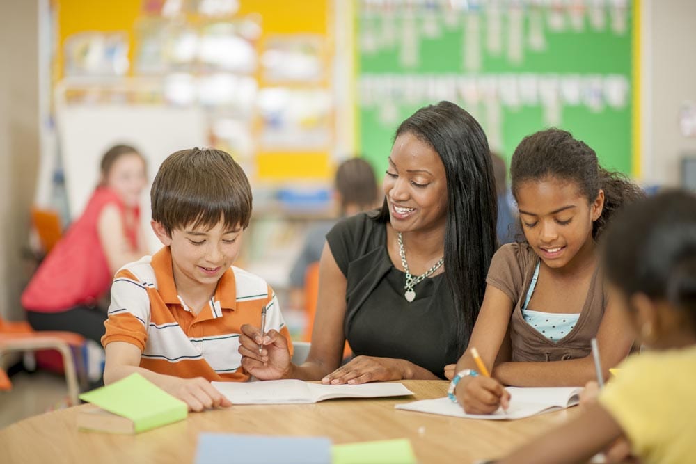 A multi-ethnic group of elementary children are sitting in class and are sitting and working on a group assignment with their teacher.