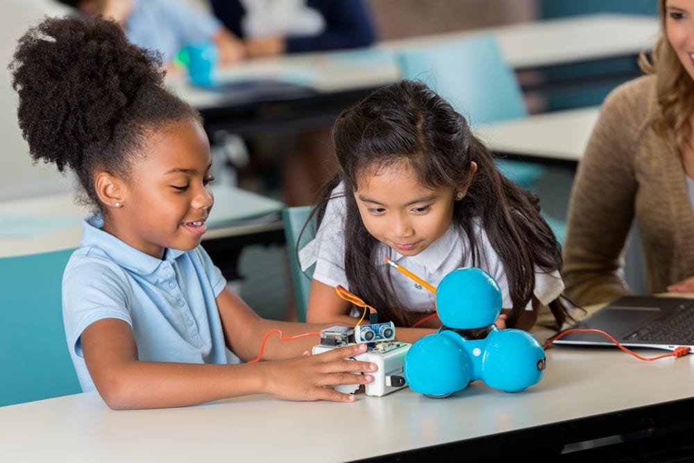 Diverse elementary schoolgirls enjoy building a robot in technology class at STEM school. A Caucasian teacher is helping them. They are wearing school uniforms.