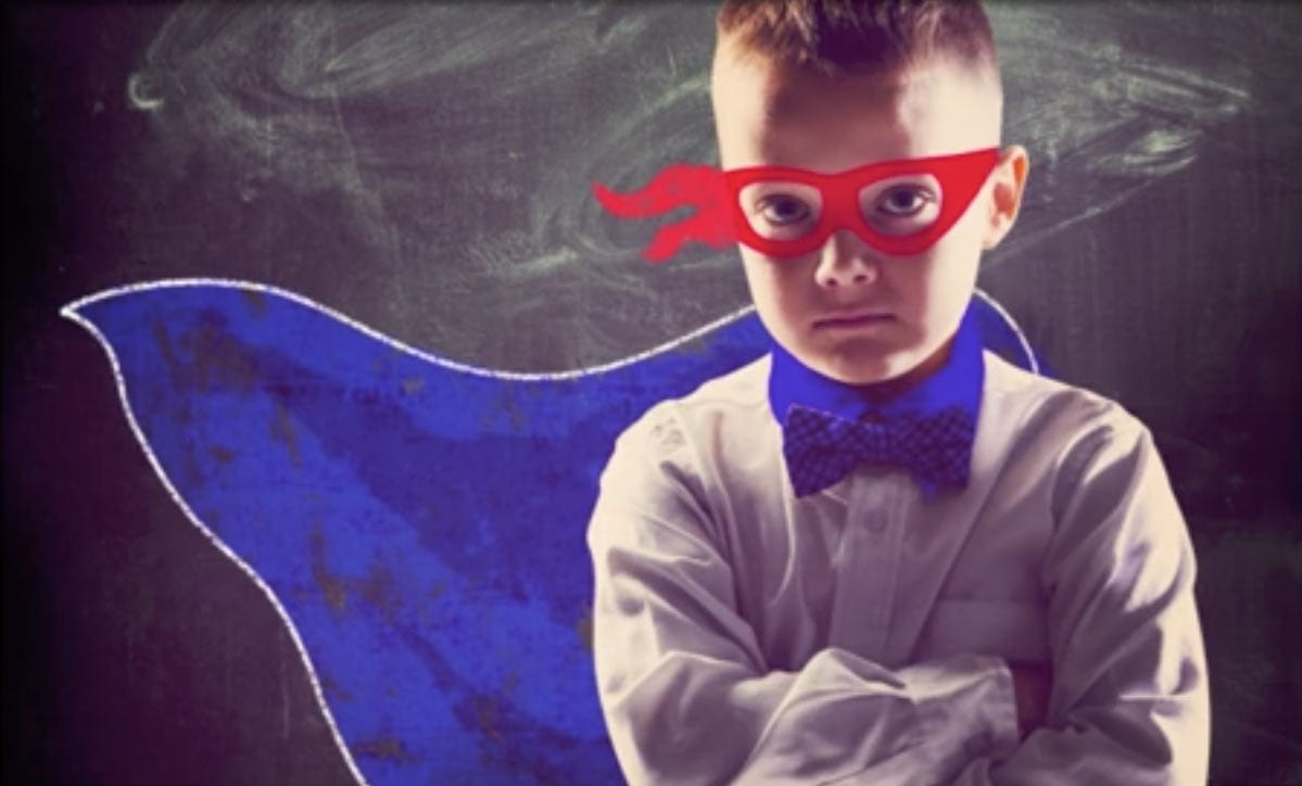 child in super hero costume with a blue cape and red mask in front of a dusty chalkboard